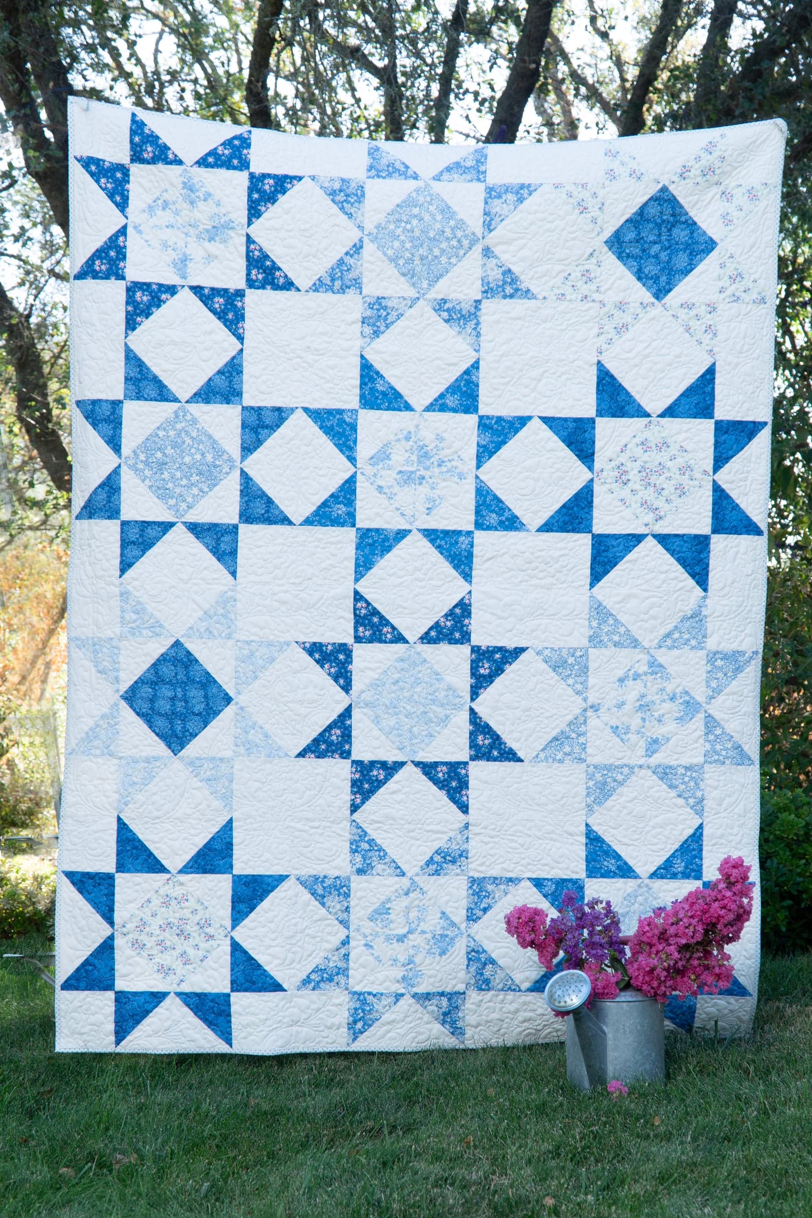 A blue and white star-block quilt hung outdoors, with a watering can of pink flowers at its foot